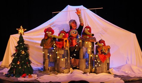 Theaterszene mit einer Person hinter vier bunten Puppen auf einer Bühne mit weißem Hintergrund und einem geschmückten Weihnachtsbaum links, © KKT. Kölner Künstler:innen Theater Theaterszene mit einer Person hinter vier bunten Puppen auf einer Bühne mit weißem Hintergrund und einem geschmückten Weihnachtsbaum links, © KKT. Kölner Künstler:innen Theater