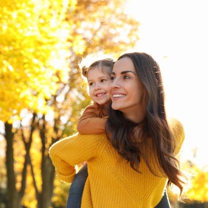 Das Bild zeigt eine Mutter mit ihrer Tochter auf dem Rücken, die draußen durch eine sonnige Herbstlandschaft laufen., © stock.adobe.com - New Africa