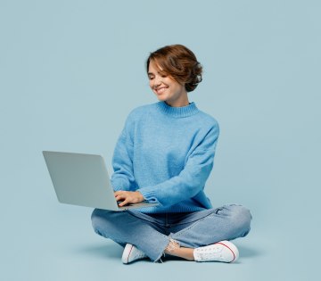Frau sitzt im Schneidersitz auf dem Boden und schaut auf das aufgeklappte Notebook, das auf ihrem Scho&szlig; liegt, &copy; Adobe Stock