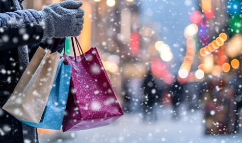 Zu sehen ist eine verschneite Ladenstraße, auf der eine Hand Einkaufstüten in das Bild hält., © stock.adobe.com - Alina 