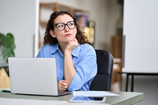 Eine junge Frau sitzt vor einem aufgeklappten Laptop und denkt nach