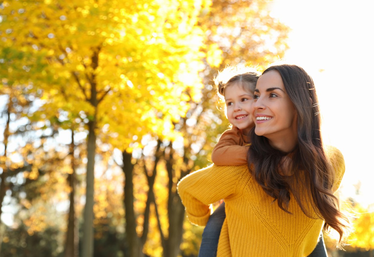 Das Bild zeigt eine Mutter mit ihrer Tochter auf dem Rücken, die draußen durch eine sonnige Herbstlandschaft laufen., © stock.adobe.com - New Africa