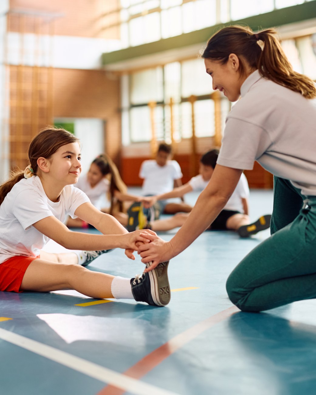 Auf dem Foto ist eine Turnhalle zu sehen. Im Vordergrund hockt eine junge Trainerin, die ein Kind dabei unterstützt sich zu dehnen., © stock.adobe.com - Drazen 