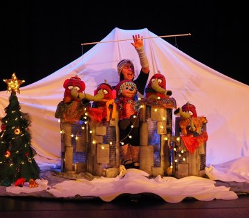 Theaterszene mit einer Person hinter vier bunten Puppen auf einer B&uuml;hne mit wei&szlig;em Hintergrund und einem geschm&uuml;ckten Weihnachtsbaum links, &copy; KKT. K&ouml;lner K&uuml;nstler:innen Theater