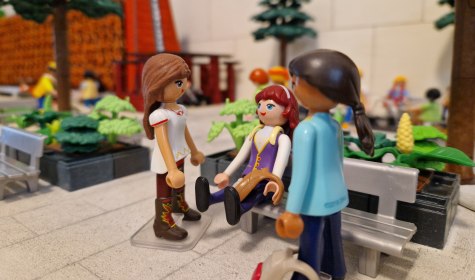 Drei Playmobil-Figuren in einem Park mit B&auml;nken und B&auml;umen, eine sitzt auf der Bank, zwei stehen davor