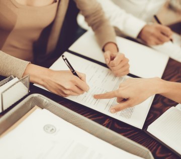 Auf dem Bild sind drei Personen zu sehen. Sie sitzen um einen Schreibtisch und haben Unterlagen auf dem Tisch ausgebreitet. Eine Person ist dabei ihre Unterschrift zu setzen. Es wird ein Vertrag &uuml;ber eine au&szlig;ergerichtliche Beilegung einer Streitigkeit aufgesetzt., &copy; VadimGuzhva - stock.adobe.com