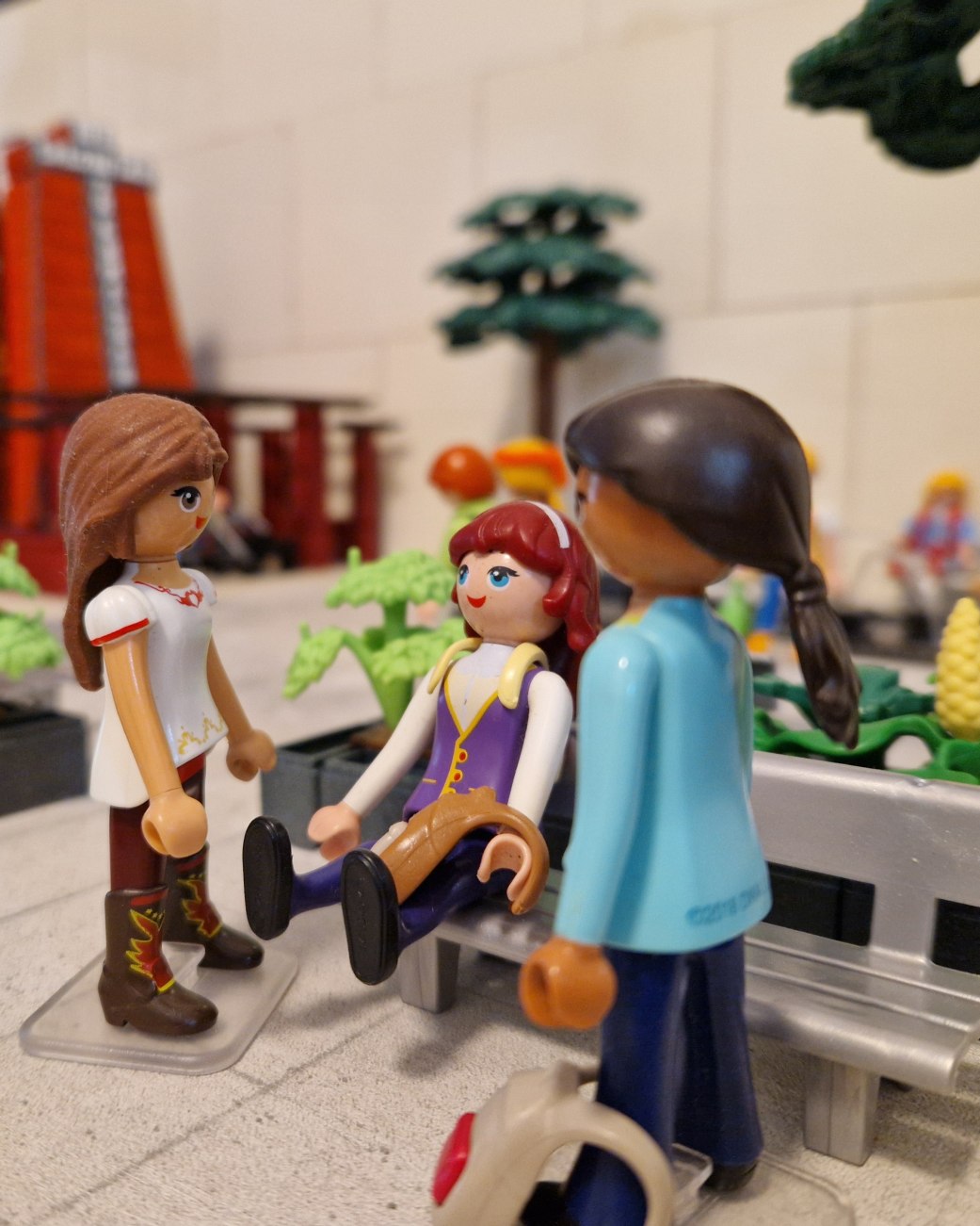 Bad Salzufler Gebäude und Bauwerke wie das Gradierwerk am Schliepsteiner Tor (Hintergrund) erwecken die Playmobil-Sammler Nicole und Michael Post im Kleinformat mit der Sonderausstellung „Nachgebaut! Bad Salzuflen aus Playmobil“ ab dem 06. November 2026 in der Wandelhalle zum Leben, © Stadt Bad Salzuflen Drei Playmobil-Figuren in einem Park mit Bänken und Bäumen, eine sitzt auf der Bank, zwei stehen davor