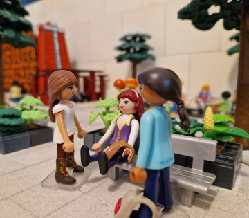 Drei Playmobil-Figuren in einem Park mit B&auml;nken und B&auml;umen, eine sitzt auf der Bank, zwei stehen davor