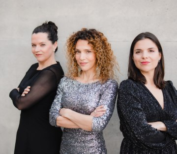 Drei Frauen mit verschr&auml;nkten Armen stehen vor einer hellen Wand, die mittlere tr&auml;gt ein glitzerndes Kleid, die anderen zwei schwarze Kleider