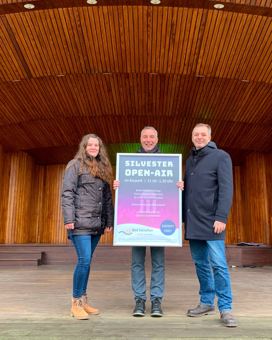 Drei Menschen stehen in der Konzertmuschel im Kurpark. In der Mitte steht Bürgermesiter Dirk Tolkemitt, er hält ein Plakat, das auf die Silvesterveranstaltung hinweist, in der Hand, © Stadt Bad Salzuflen