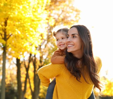 Das Bild zeigt eine Mutter mit ihrer Tochter auf dem Rücken, die draußen durch eine sonnige Herbstlandschaft laufen., © stock.adobe.com - New Africa