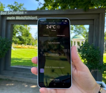 Person h&auml;lt Smartphone mit dem Startbildschirm der MeinS App in der Hand. Im Hintergrund sind der Leopoldsprudel und der Kurpark der Stadt Bad Salzuflen zu sehen, &copy; Stadt Bad Salzuflen