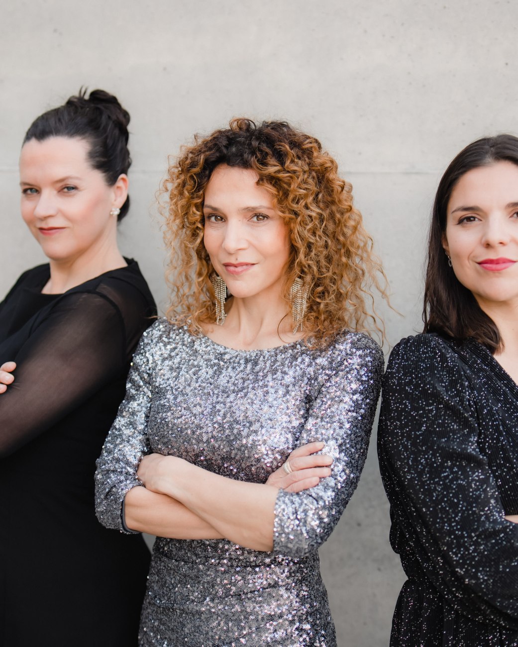 Drei Frauen mit verschr&auml;nkten Armen stehen vor einer hellen Wand, die mittlere tr&auml;gt ein glitzerndes Kleid, die anderen zwei schwarze Kleider