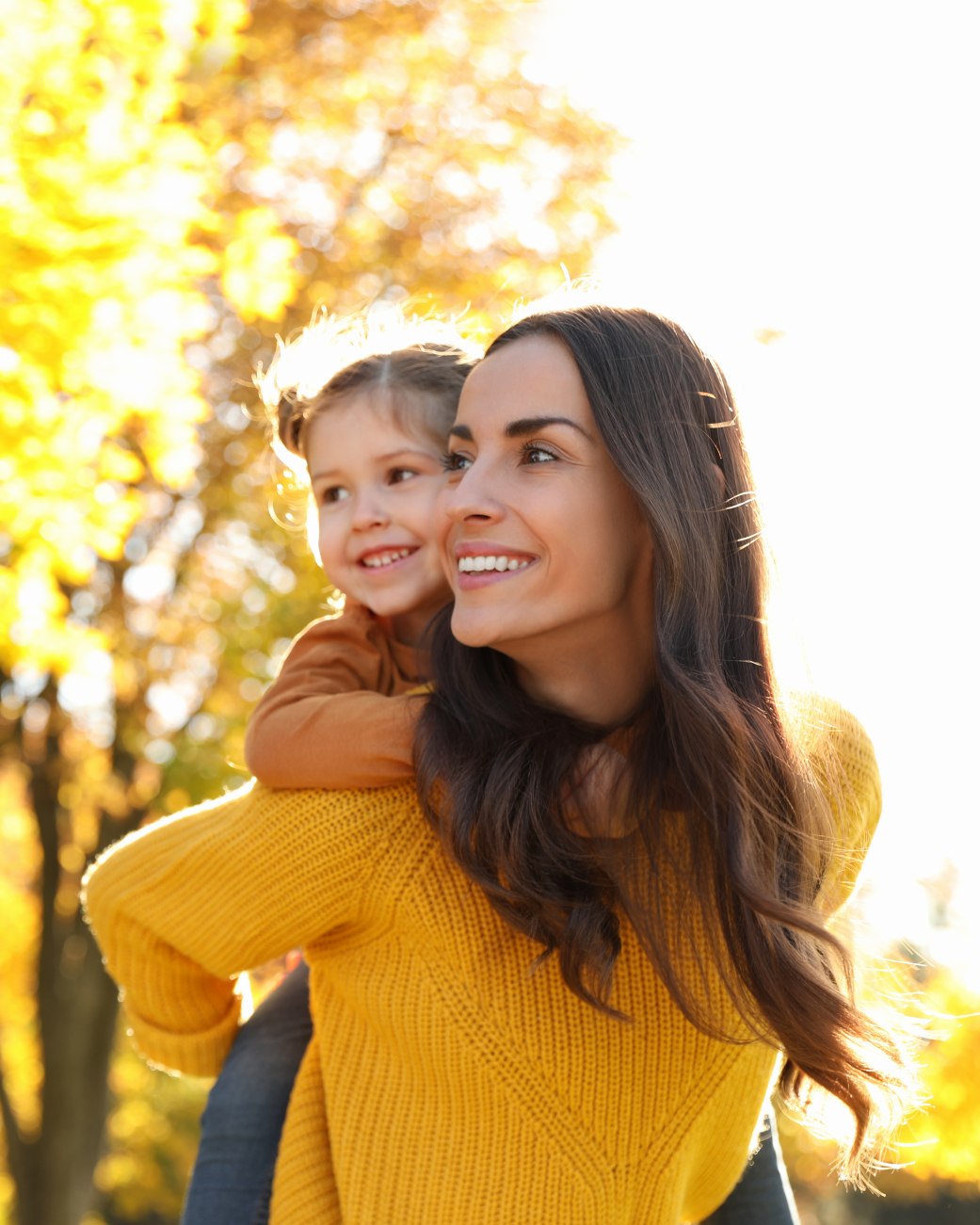 Das Bild zeigt eine Mutter mit ihrer Tochter auf dem Rücken, die draußen durch eine sonnige Herbstlandschaft laufen., © stock.adobe.com - New Africa