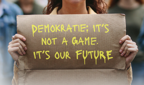 Auf dem Bild ist eine weiblich gelesene Person zu sehen, die ein Plakat vor sich hoch hält auf dem &quot;Demokratie: it's not a game, it's our future!&quot; geschrieben steht., © Stock.adobe.com - Jacob Lund