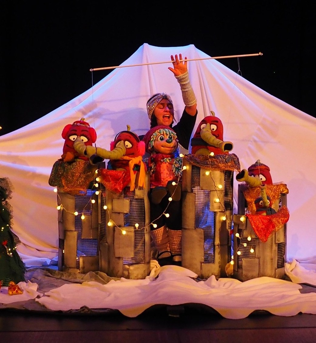Theaterszene mit einer Person hinter vier bunten Puppen auf einer B&uuml;hne mit wei&szlig;em Hintergrund und einem geschm&uuml;ckten Weihnachtsbaum links, &copy; KKT. K&ouml;lner K&uuml;nstler:innen Theater