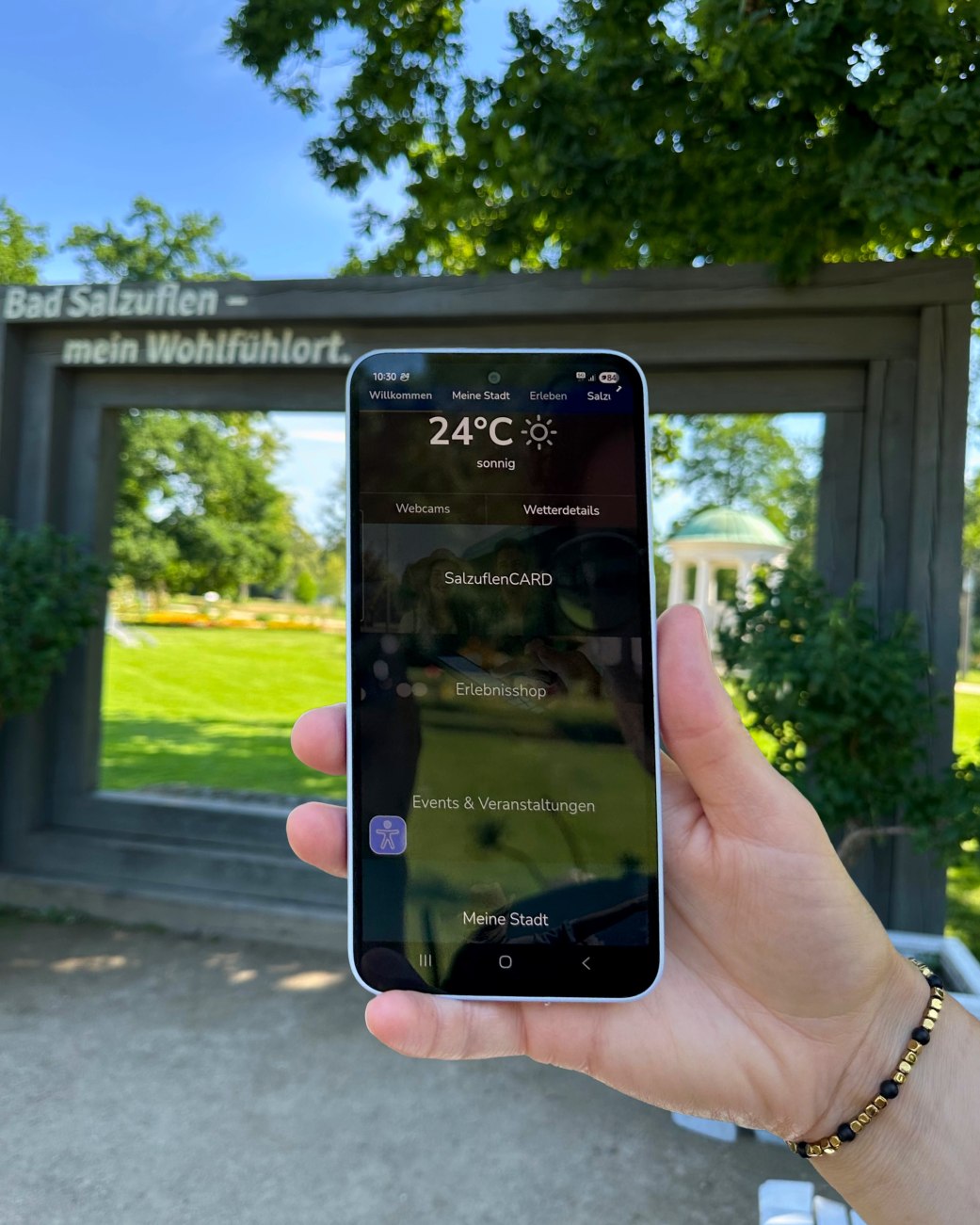 Person hält Smartphone mit dem Startbildschirm der MeinS App in der Hand. Im Hintergrund sind der Leopoldsprudel und der Kurpark der Stadt Bad Salzuflen zu sehen, © Stadt Bad Salzuflen Person hält Smartphone mit dem Startbildschirm der MeinS App in der Hand. Im Hintergrund sind der Leopoldsprudel und der Kurpark der Stadt Bad Salzuflen zu sehen, © Stadt Bad Salzuflen
