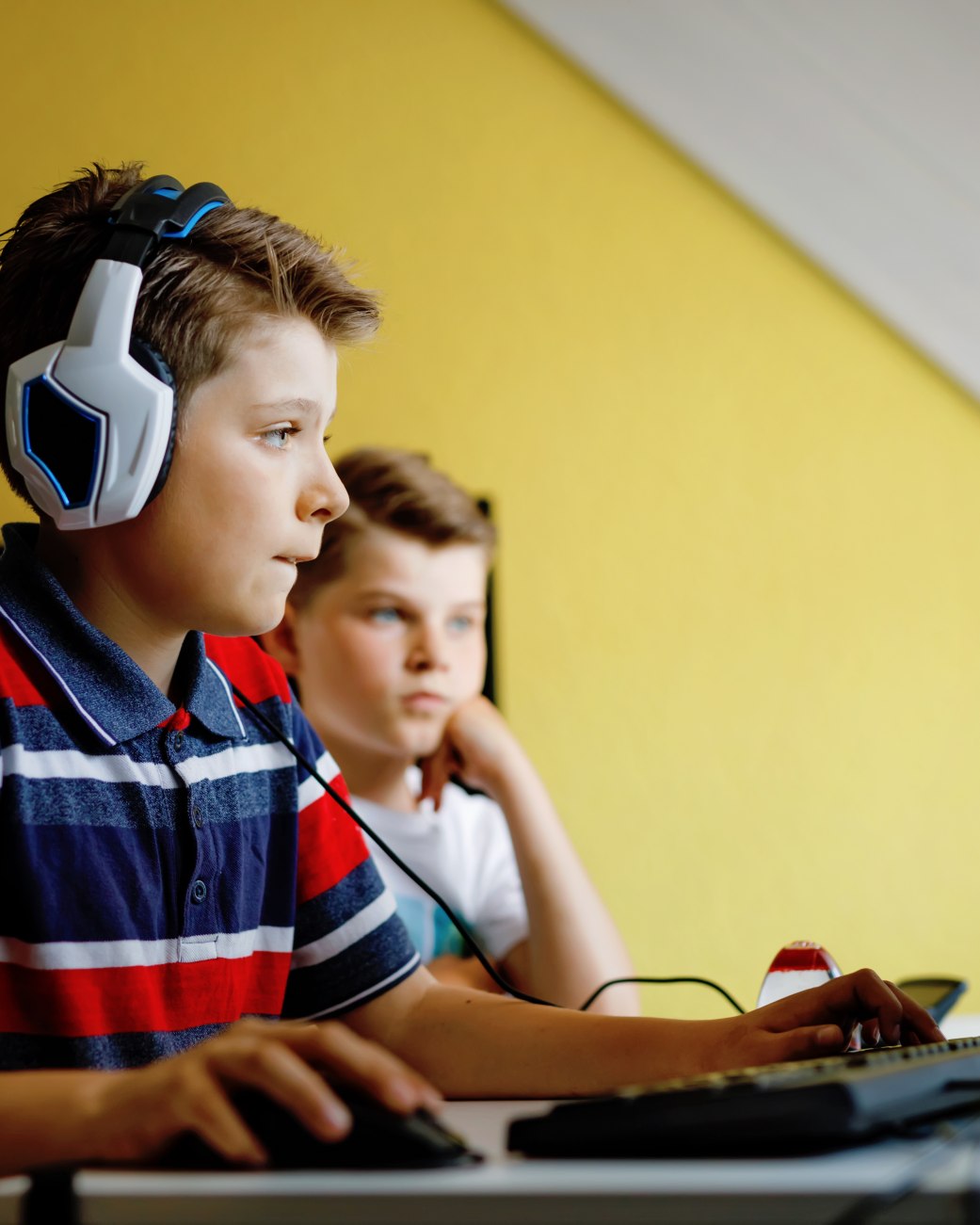 Ein Kind sitzt mit Kopfhörern vor einem PC, ein anderes Kind schaut zu, © stock.adobe.com - Irina Schmidt
