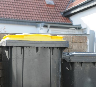 Auf dem Bild zu sehen ist eine Mauer, vor der verschiedene M&uuml;lltonnen stehen. Im Mittelpunkt des Bildes steht eine schwarze M&uuml;lltonne mit gelbem Deckel. Es handelt sich um die Gelbe Tonne f&uuml;r Leichtverpackungen., &copy; fotoak80 - stock.adobe.com